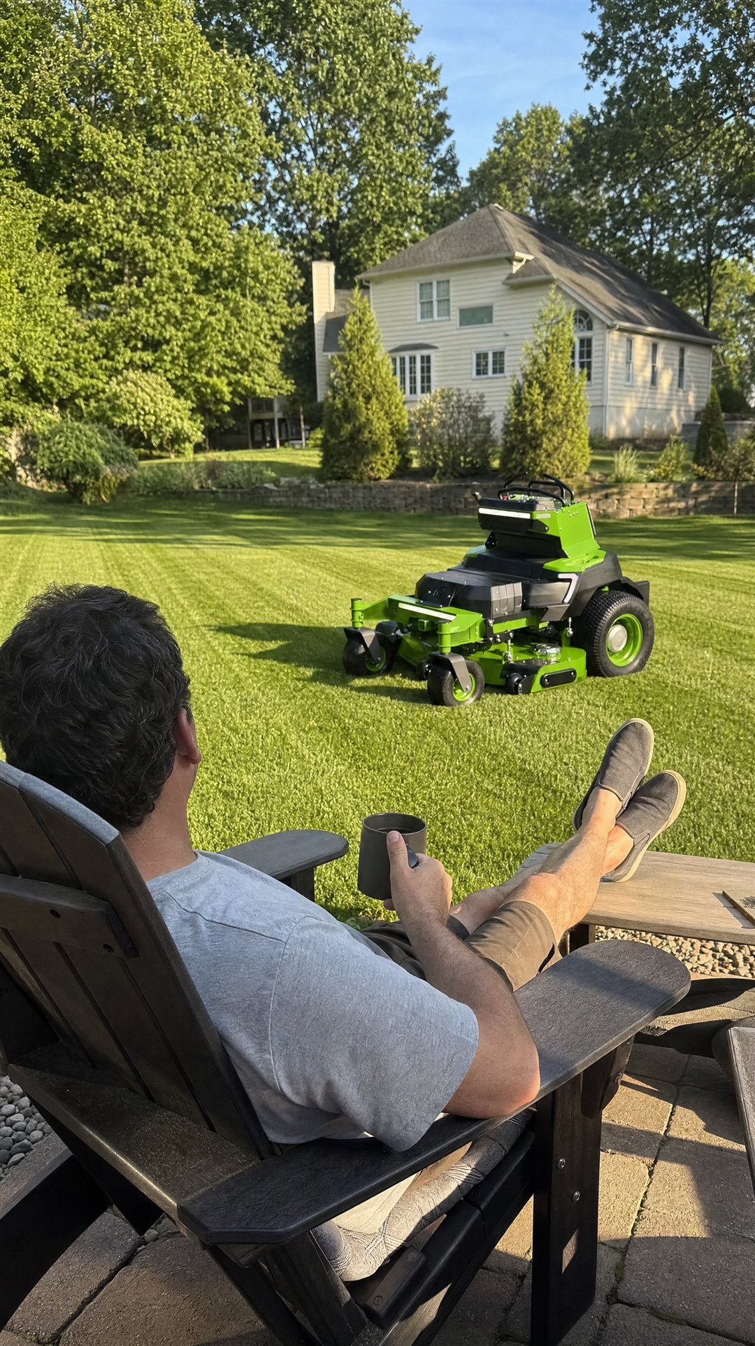 A homeowner relaxing with coffee while the quiet all-electric mower finishes a freshly striped lawn